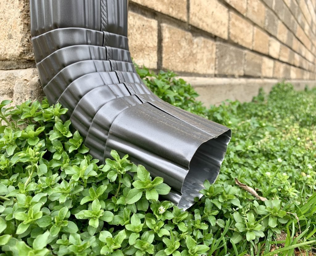 Close-up of a black metal downspout draining water into green plants near a brick wall, installed by The Exterior Zone in Lake Orion, Michigan.